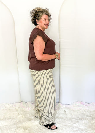 A woman modeling a brown sleeveless top and beige-and-green vertically striped pants, standing in front of a white arched backdrop.