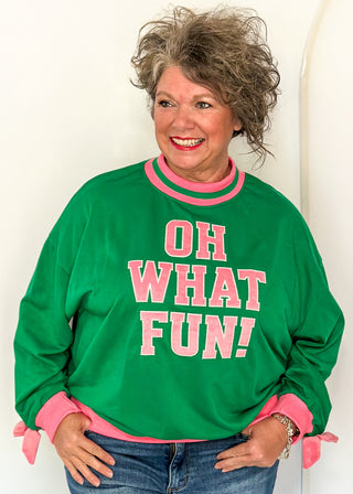 woman wearing a green long sleeve mock neck top with pink contrast sleeve  and "OH WHAT FUN" embroidered letter patches.