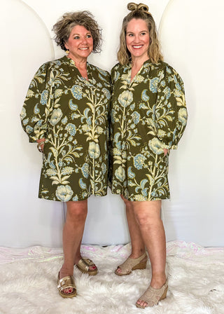 Two women modeling matching olive green floral print mini robes with blue and beige details, standing on a white faux fur rug against a white studio backdrop.