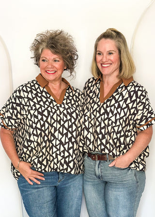 Two women standing side-by-side wearing matching short-sleeve patterned tops with a brown v-neck collar, paired with blue jeans.