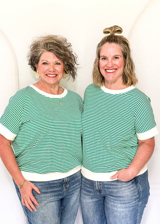 Two women wearing matching green and white striped shirts and blue jeans against a white background.