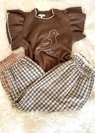 A children's two-piece outfit laid flat on a white faux fur background. The set includes a brown short-sleeve top with ruffled sleeves and a white bird outline on the chest, paired with brown and beige plaid pants featuring a solid brown side panel.