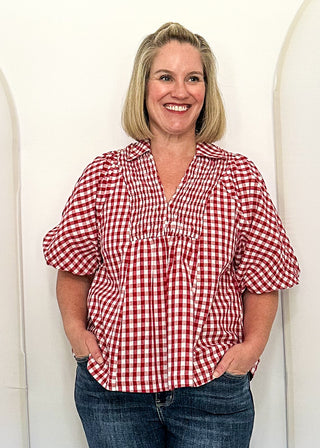 A smiling blonde woman stands indoors between three large white arched panels, wearing a red-and-white gingham short-sleeve blouse and dark blue jeans.