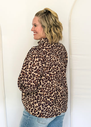 A woman with blonde hair tied back is shown from the side/back wearing a brown and beige leopard print long-sleeve shirt and blue jeans, standing against a white studio backdrop.