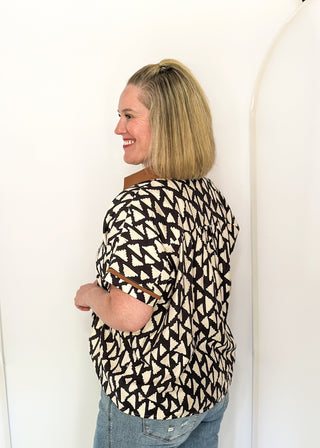 A woman with blonde hair is shown from the side, wearing a short-sleeve top with a black and cream geometric pattern and light blue jeans, standing against a white wall.
