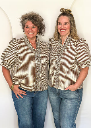 Two women standing side-by-side wearing matching beige and brown vertically striped short-sleeve button-up shirts with ruffle trim, paired with blue jeans, against a white arched backdrop.