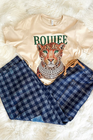 A beige graphic T-shirt with a leopard print and the word 'BOUIEE' in green, paired with blue and navy plaid shorts, laid flat on a white faux fur background.