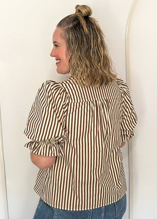 A woman with blonde hair in a bun is shown from the back wearing a brown and beige vertically striped blouse with ruffled sleeves, paired with blue jeans, standing against a white studio backdrop with arch-shaped props.