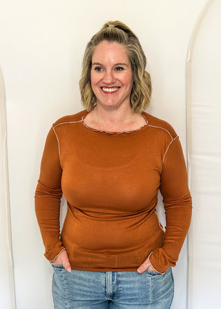 A smiling woman with blonde hair tied back stands against a white arched backdrop, wearing a fitted rust-orange long-sleeve top and light blue jeans.