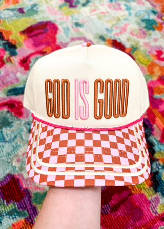 A cream-colored baseball cap held by a hand, featuring 'GOD IS GOOD' embroidered on the front in brown and pink, with a pink and cream checkered pattern on the visor. The background is a colorful, textured blanket.