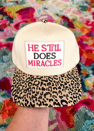A baseball cap held by a hand, featuring an off-white crown with a white patch that reads 'HE STIL DOES MIRACLES' in red and black text, and a leopard-print brim. The background is a colorful, textured blanket.