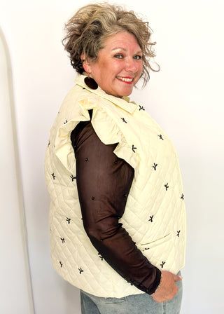 Ivory quilted puffer vest-ruffle sleeve, front pockets, snap closure. Small black bows embroidered throughout.  