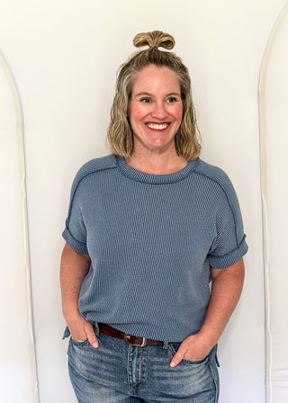 Woman wearing a blue sweater and jeans standing against a white wall.