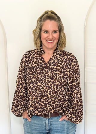 A smiling woman with blonde hair stands against a white arched backdrop, wearing a leopard-print long-sleeve blouse with blue jeans.