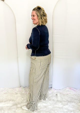 A woman stands in profile wearing a dark navy long-sleeve top and beige pinstriped pants with a drawstring waist, standing on a white faux fur rug in front of white arch backdrops.