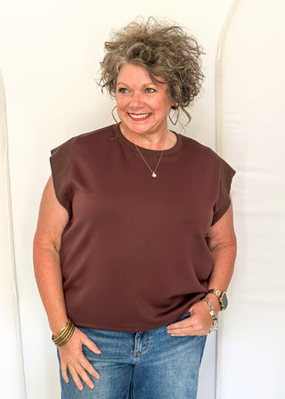 A smiling woman with curly gray hair wearing a sleeveless, cropped brown top and blue jeans, standing against a white arched backdrop.