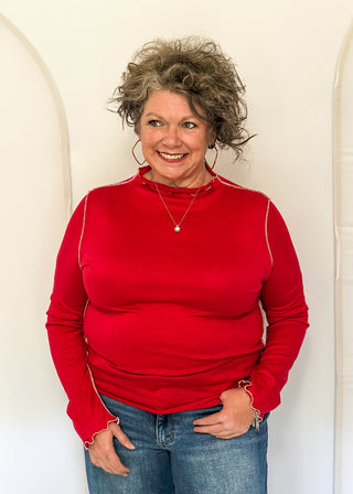 A smiling woman with curly gray hair wearing a bright red long-sleeve mock neck top and blue jeans, standing against a white arched backdrop.