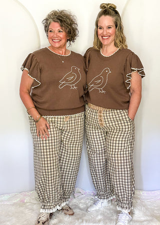 Two women modeling matching brown short-sleeve t-shirts with white bird embroidery and beige-and-white gingham pants, standing against a white studio backdrop on a white faux-fur rug.