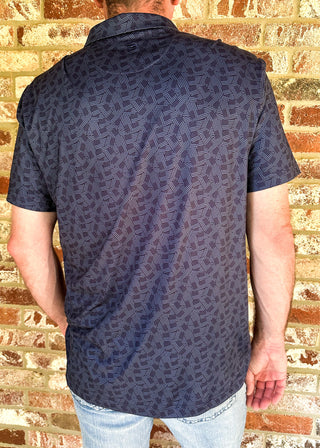 A man viewed from behind wearing a short-sleeve button-up shirt with a dark navy blue and black geometric pattern, paired with light blue jeans, standing against a brick wall.