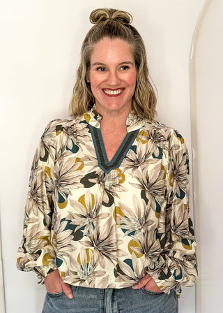 A smiling woman with blonde hair in a bun stands against a white arched wall, wearing a cream-colored long-sleeve blouse with a tropical floral print in green, mustard yellow, and brown, paired with blue jeans.