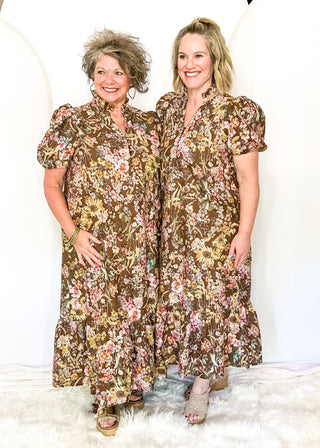 Two women standing side by side wearing matching brown floral midi dresses with puff sleeves, posed against a white studio backdrop with a white faux fur rug.