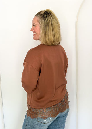 A woman with blonde hair tied back is shown from the back, wearing a rust-brown crewneck sweatshirt, a brown lace hem detail, and blue jeans against a white arched wall.
