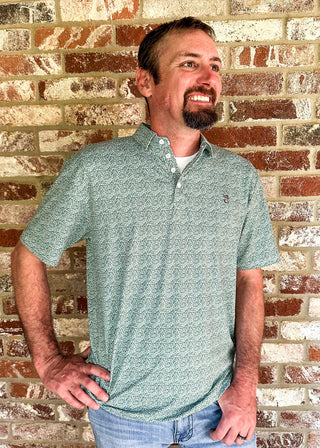 A man wearing a sage green short-sleeve polo shirt with a subtle geometric pattern, standing against a rustic red and beige brick wall.
