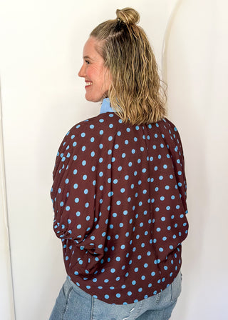 A woman shown from the back wearing a brown long-sleeve button-up shirt with light blue polka dots, paired with light-wash blue jeans, standing against a white studio backdrop with arched panels.