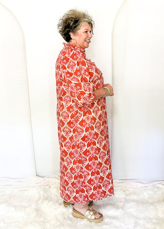 A woman wearing a red and white floral print midi dress, standing in profile against a white studio backdrop with arched panels.