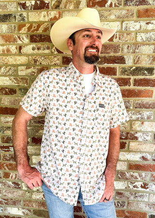 A man wearing a cream cowboy hat, a short-sleeve button-up shirt with a white background and a green and orange allover pattern, and blue jeans, standing against a rustic red and beige brick wall.