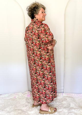 An older woman stands in profile wearing a long-sleeved, high-neck maxi dress with a dense red, pink, and beige floral pattern on a dark background. She is standing on a white faux fur rug in front of a white arched backdrop.