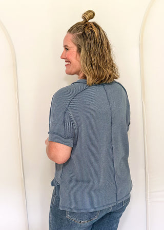 Person wearing a blue ribbed sweater and jeans standing against a white wall.