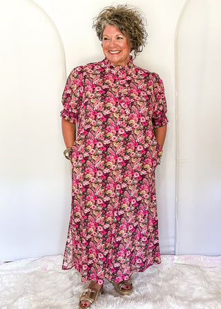 A woman modeling a pink and red floral midi dress with pockets and elbow-length sleeves, standing on a white faux fur rug against a white arched backdrop.