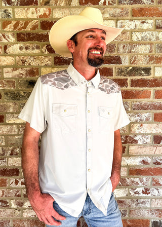 A man wearing a white short-sleeve button-up shirt with gray patterned shoulder yokes, a cream cowboy hat, and blue jeans, standing against a rustic brick wall.