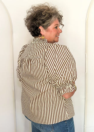A woman with short curly gray hair is shown from the back wearing a brown and beige vertically striped blouse with puffed sleeves and blue jeans, standing against a white arched studio backdrop.