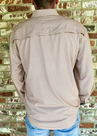 A man viewed from behind wearing a light beige long-sleeve button-down shirt with a subtle grid pattern, paired with blue jeans and a beige baseball cap, standing against a rustic brick wall.