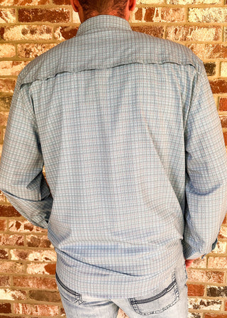 A man viewed from behind wearing a light blue and white plaid western-style long-sleeve shirt and light blue jeans, standing against a rustic red-brown brick wall.