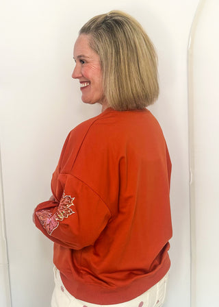side view of woman wearing an orange long sleeve top with sequin leaves on the sleeves.