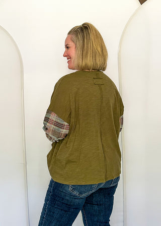 A woman with blonde hair is shown from the back wearing an olive green top with plaid elbow patches and blue jeans, standing in front of a white arch-shaped backdrop.