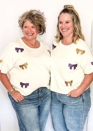 Short sleeve white top featuring scattered sequin bows in gold and purple