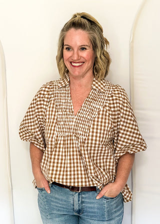 A smiling woman with blonde hair tied back stands against a white arched backdrop, wearing a brown and beige gingham short-sleeve blouse with blue jeans.