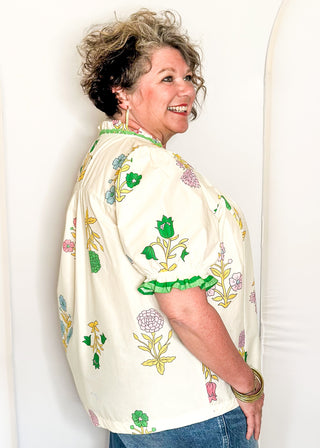 off white floral short sleeve button front blouse with pink and green flowers, featuring puff sleeves, ruffled neckline, and green trim.