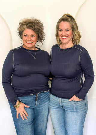 Two women standing side-by-side wearing matching dark navy long-sleeve crewneck tops and blue jeans, posing against a white arched studio backdrop.