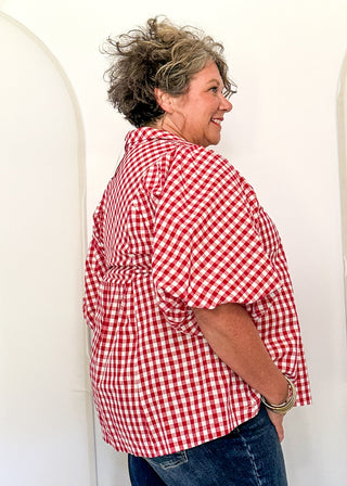 A woman wearing a red and white gingham short-sleeve button-up shirt and blue jeans, standing in front of a white arched backdrop.