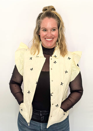 Ivory quilted puffer vest-ruffle sleeve, front pockets, snap closure. Small black bows embroidered throughout.  