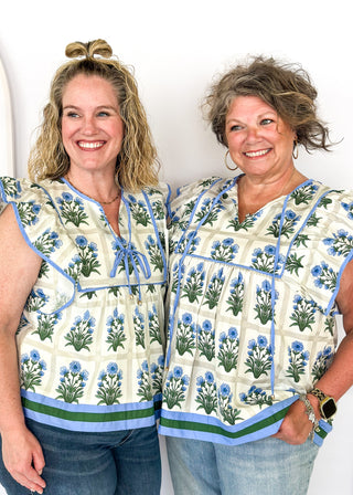 Flutter sleeve top with off white background and gray grid pattern-large blue florals with green stems/leaves in each grid.  Blue ribbon trim on sleeve and collar.  Blue ribbon tie neck.  Blue and dark green trim on bottom band. 