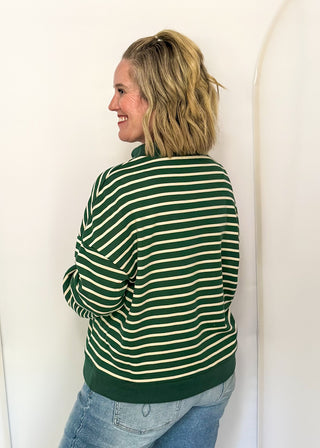 A woman with blonde hair tied back is shown from the back wearing a green and cream striped sweater and blue jeans, standing against a white studio backdrop.