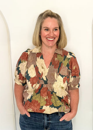 A smiling blonde woman wearing a short-sleeve button-up blouse with a brown, beige, and green floral pattern, paired with blue jeans, standing against a white arched backdrop.