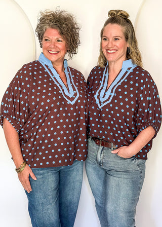 Two women modeling matching brown and light blue polka dot tops with light blue v-neck trim, paired with blue jeans, standing against a white arched backdrop.