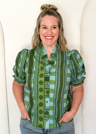 A woman with blonde hair in a bun stands smiling, wearing a green and light blue patterned short-sleeve blouse with puff sleeves and blue jeans, set against a white arched wall.
